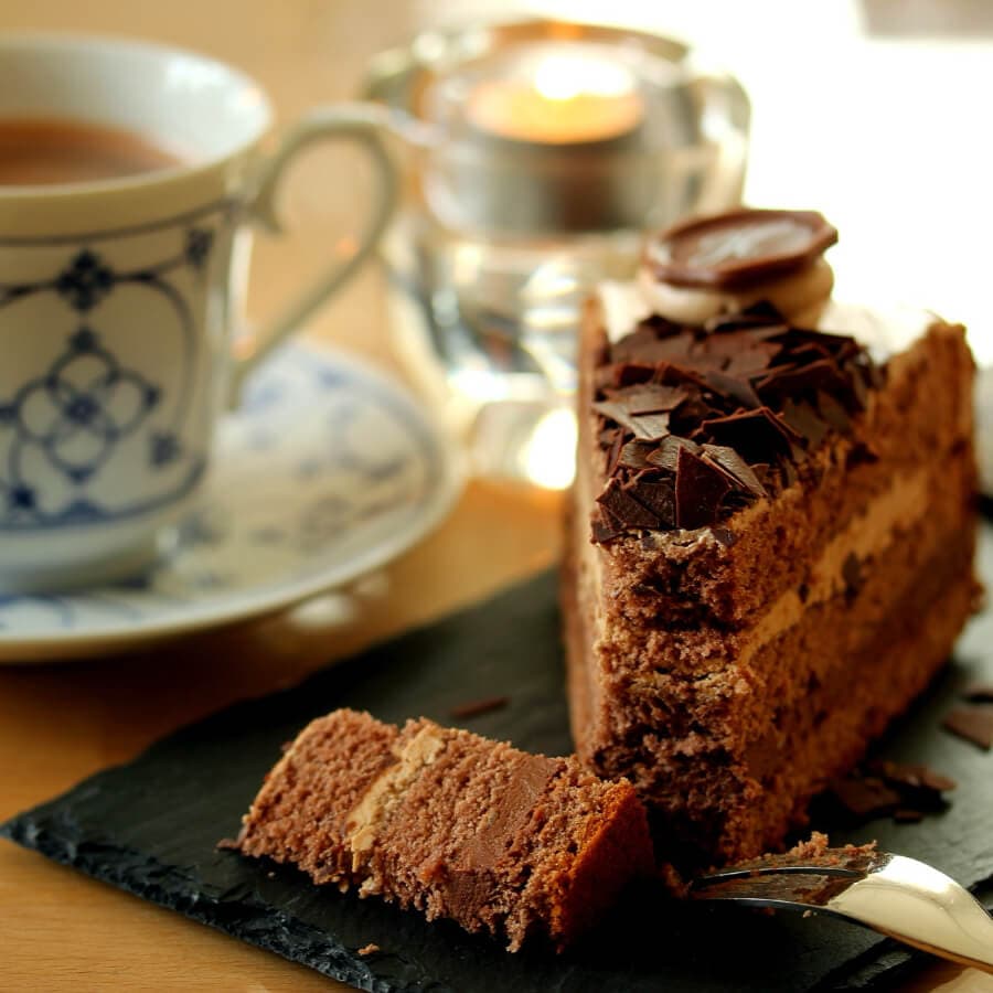 A slice of chocolate cake on a black slate, accompanied by a cup of tea and a flickering candle.