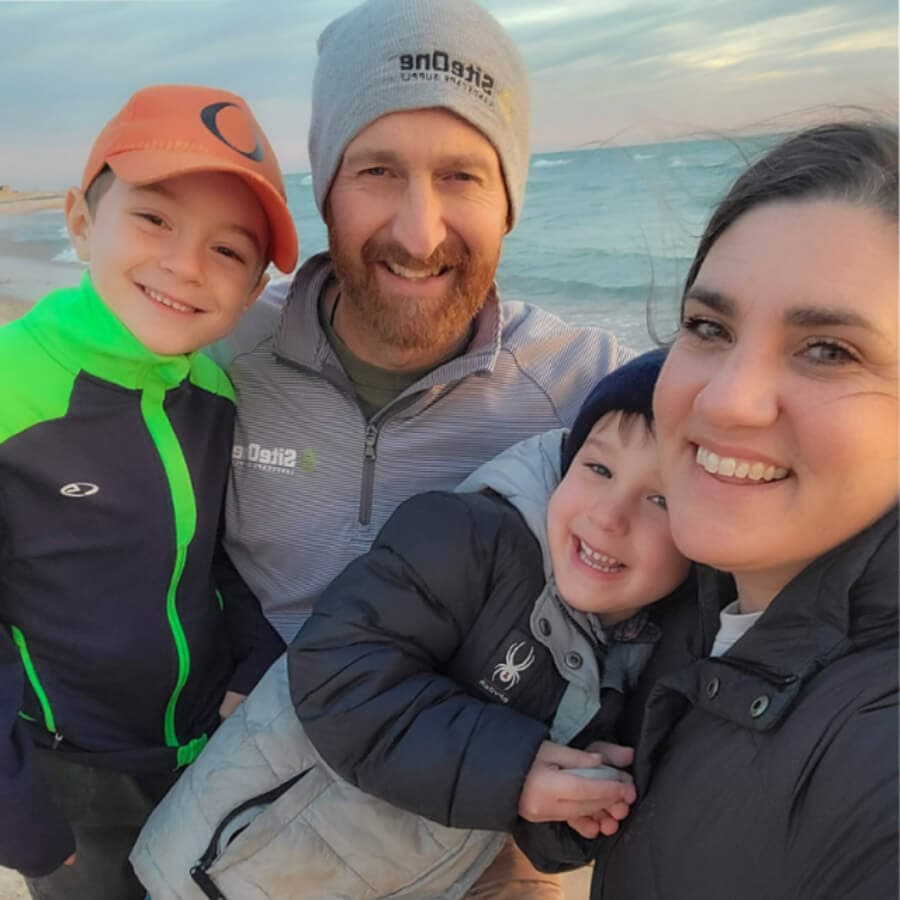 A smiling family of four poses together on the beach during sunset.