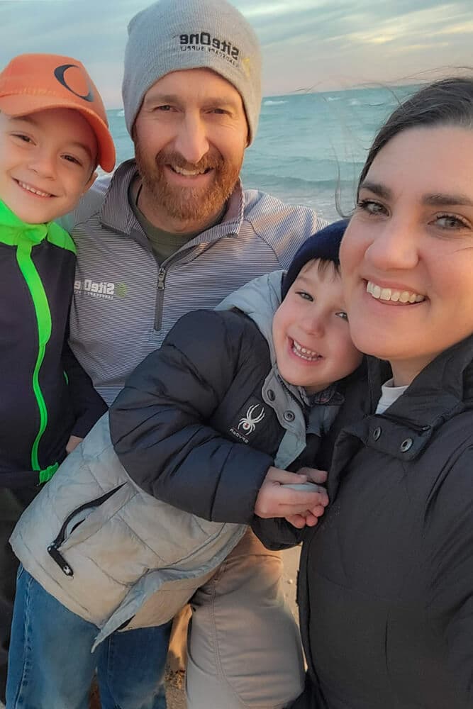 A smiling family with two young boys poses for a selfie at the beach during sunset.