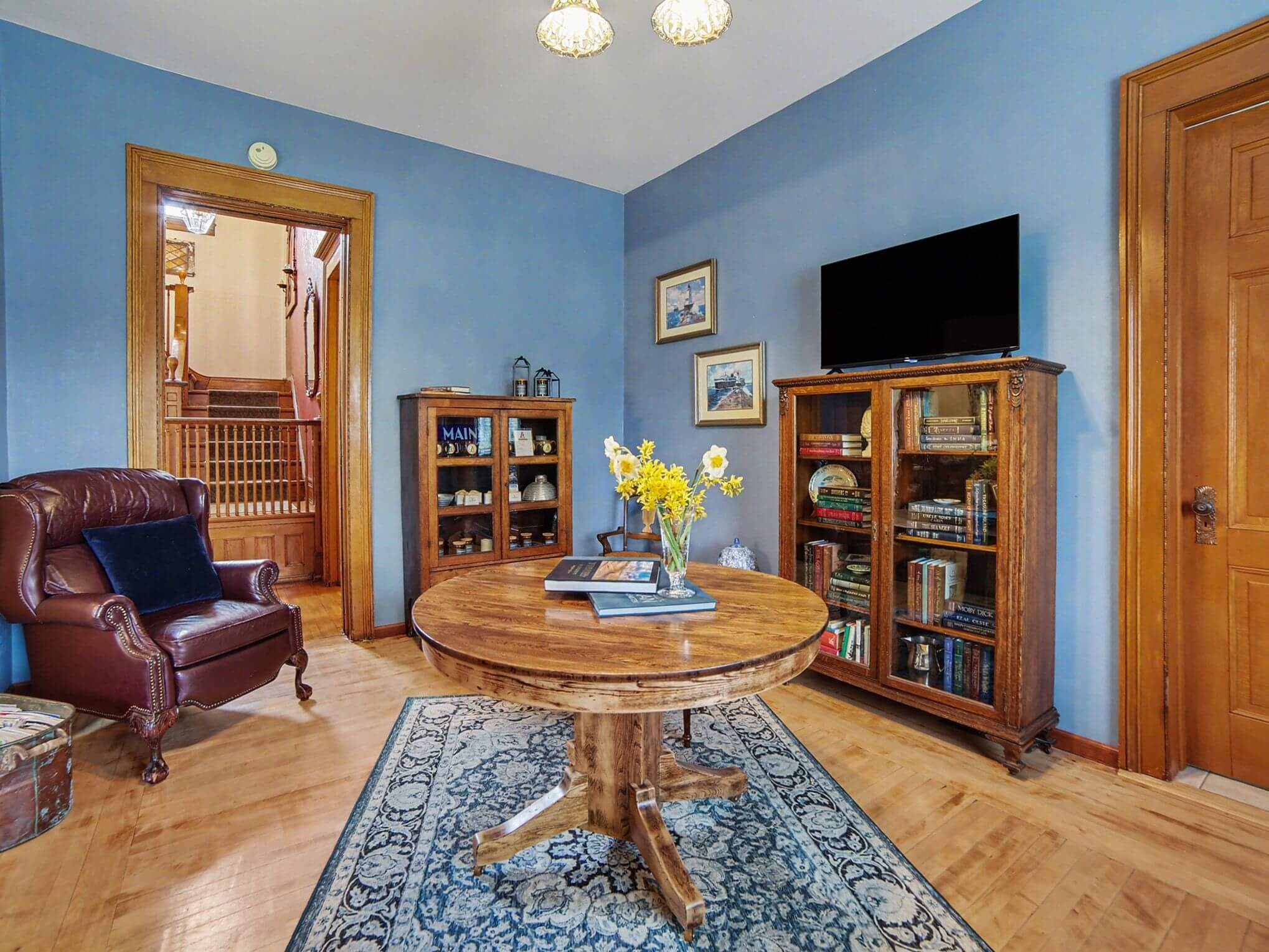A cozy blue-walled room featuring a wooden table, bookshelves, a TV, and a leather armchair.