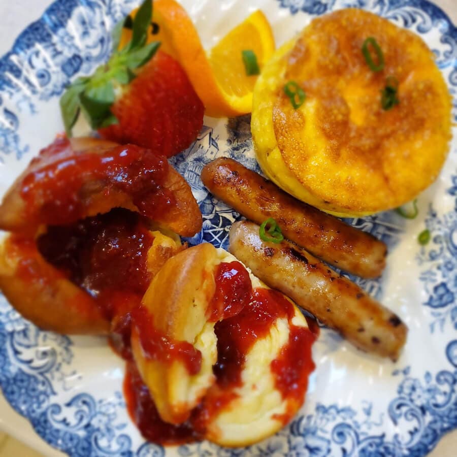 A plate of breakfast featuring sausages, a fluffy omelet, and strawberry jam-filled pastries, garnished with fruit slices.