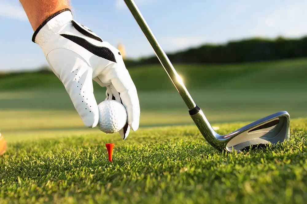 A golfer in a white glove places a golf ball on a tee in the grass.