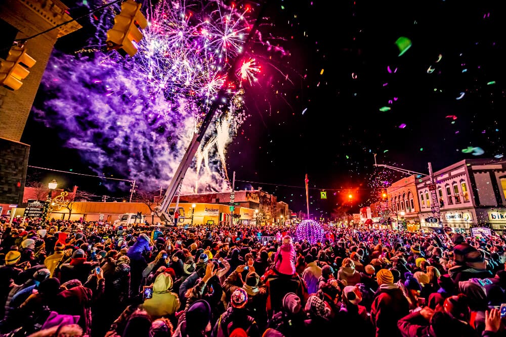 A large crowd celebrates under vibrant fireworks and confetti during a nighttime event in a bustling town.