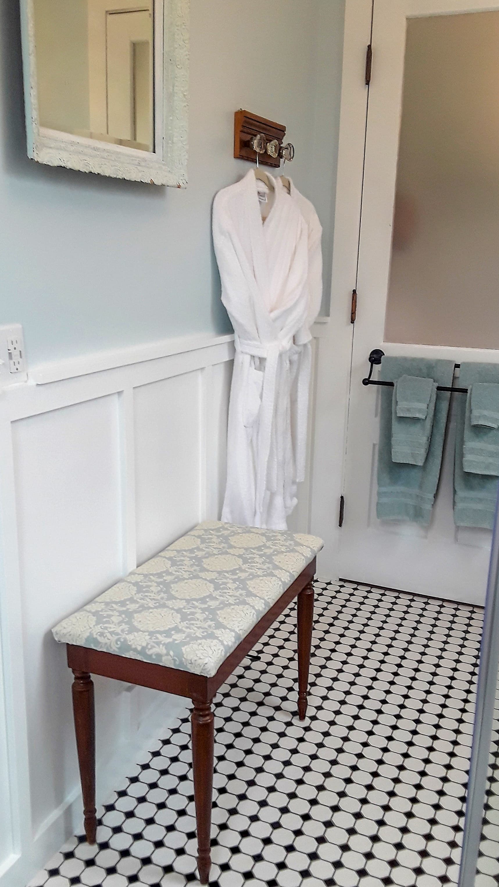 A cozy bathroom featuring a white robe on a hook, a patterned bench, and teal towels.