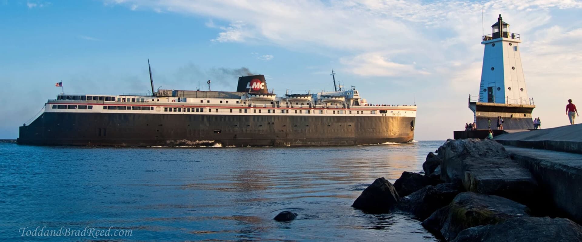 A large ship sails past a lighthouse along a rocky shoreline under a blue sky.