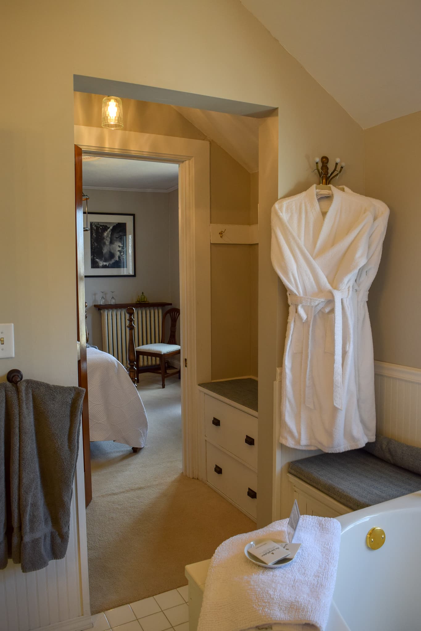 A cozy bathroom with a plush white robe hanging and a view into a softly lit bedroom.