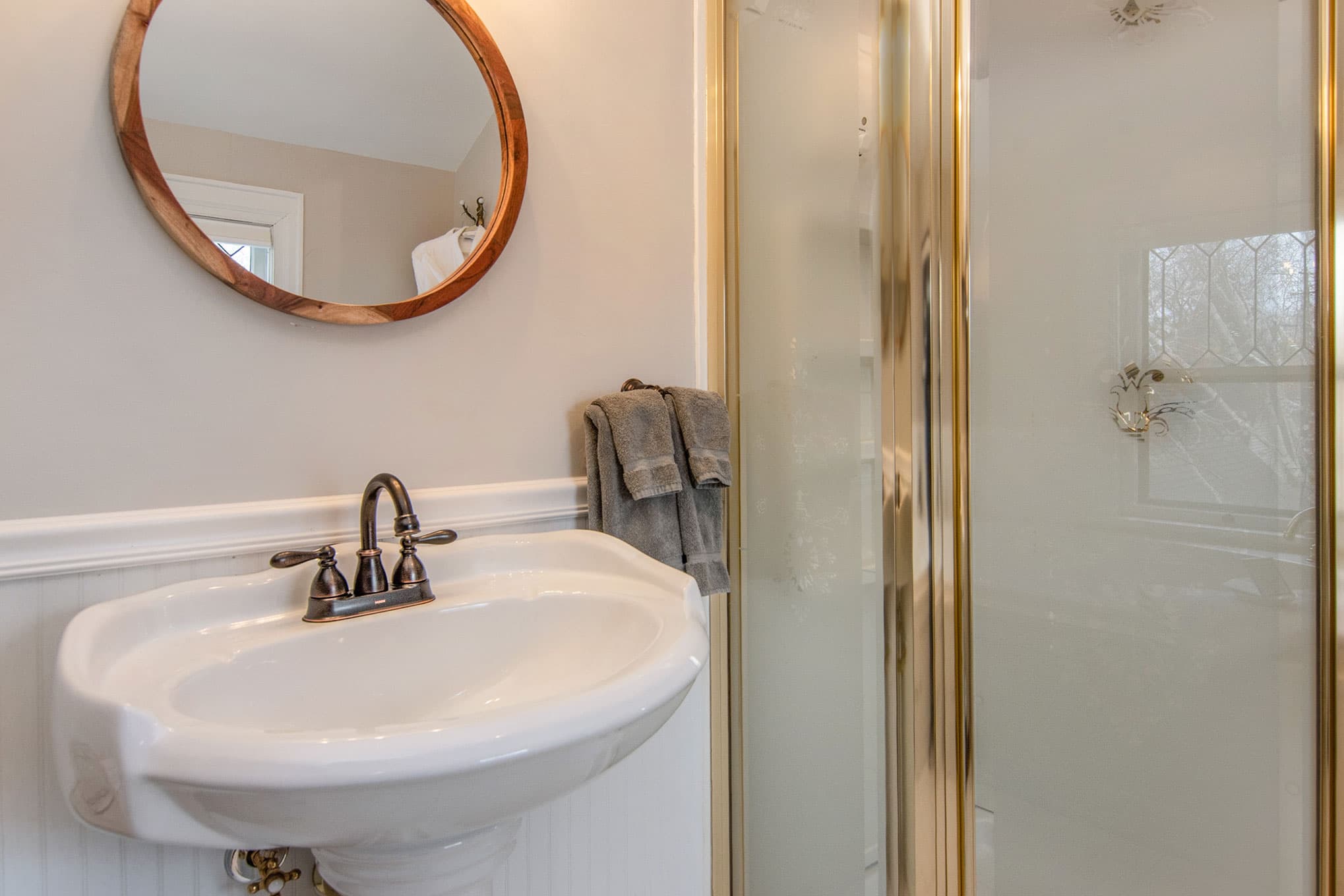 A modern bathroom featuring a pedestal sink, a round mirror, and a glass shower enclosure.