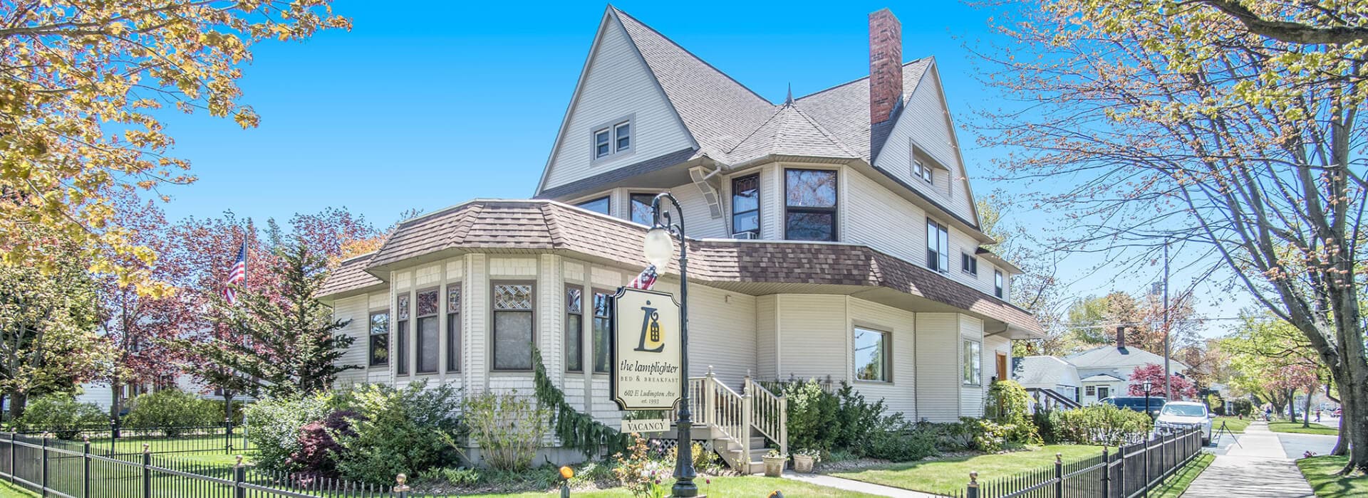 A charming, two-story house with a gabled roof and a front porch, surrounded by greenery and blooming trees.
