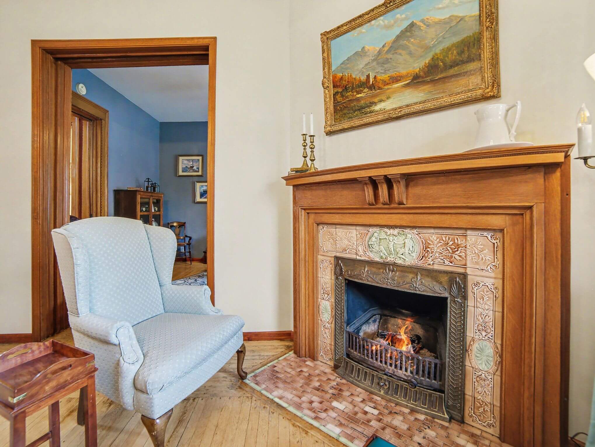 Cozy living room featuring a blue armchair beside a fireplace with a decorative mantel and a landscape painting above.