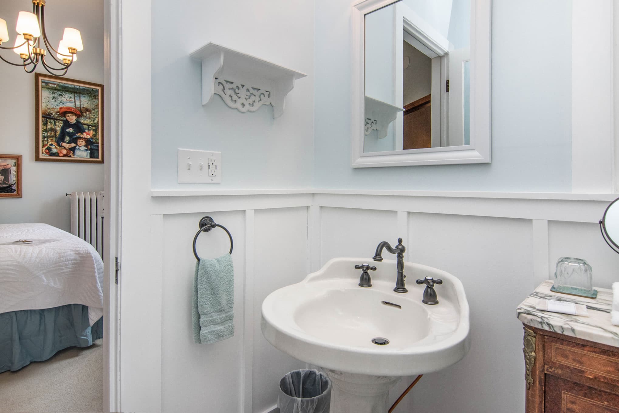 A small bathroom featuring a vintage sink, light blue walls, and a mirror, with a towel and artwork in the background.