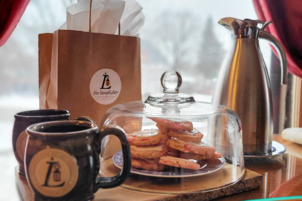 A cozy display featuring cookies under a glass dome, two mugs, a coffee pot, and a paper bag from a café.