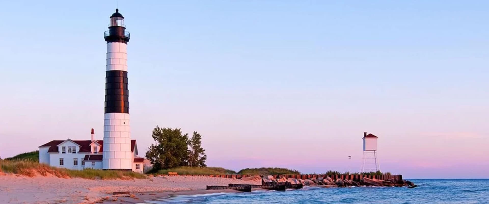 A black and white striped lighthouse beside a calm beach at sunset.