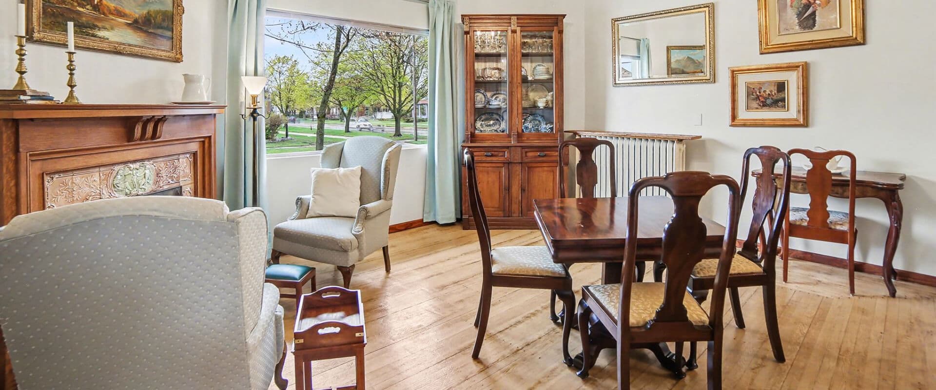 Cozy living room featuring a vintage dining set, armchairs, and a large window overlooking greenery.