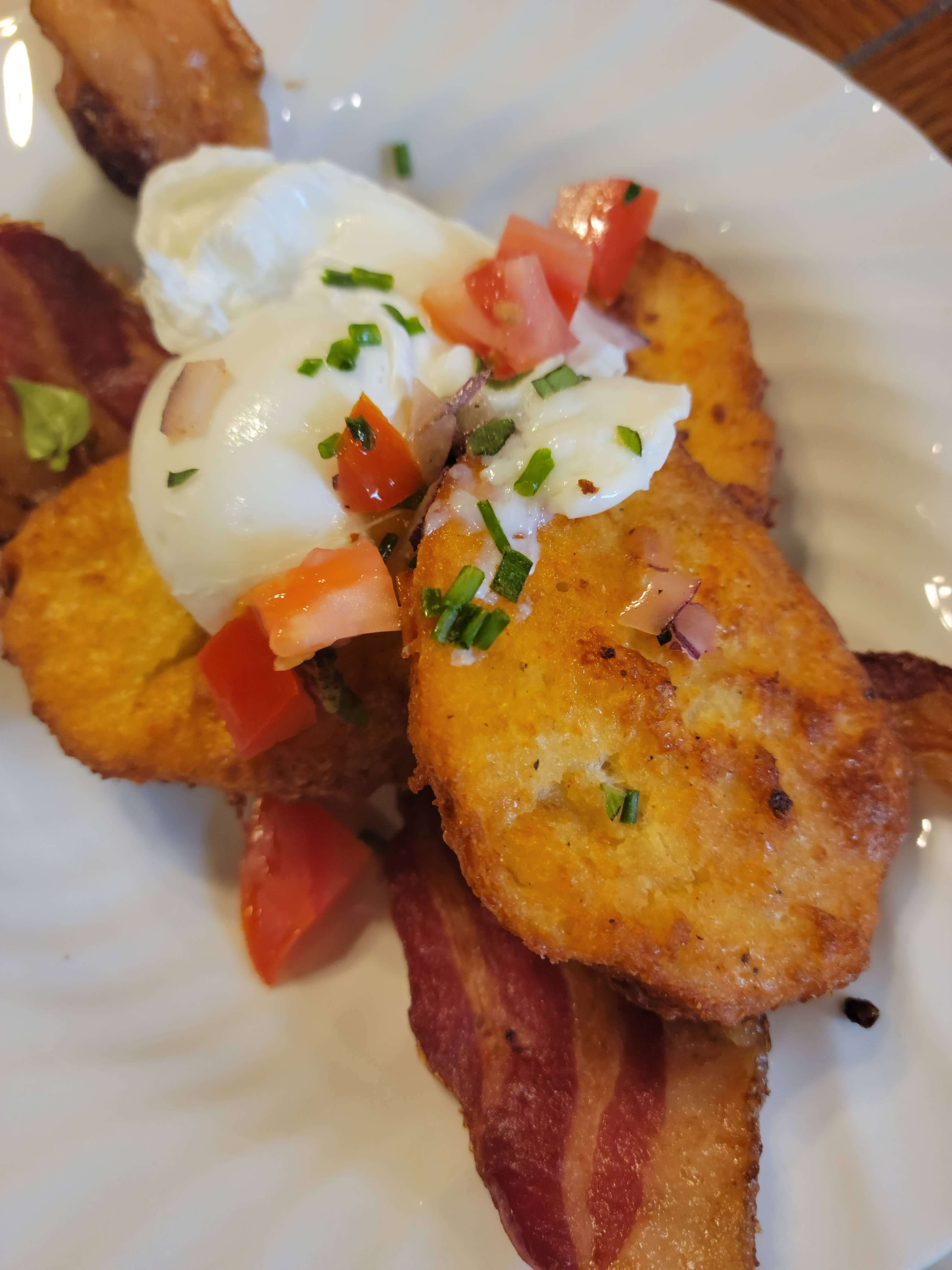 A close-up of crispy potato cakes topped with poached egg, diced tomatoes, and chives, served with crispy bacon.