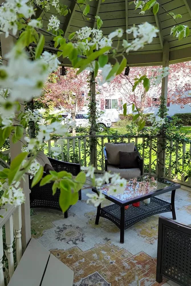 A cozy gazebo furnished with seating and a glass table, surrounded by blooming flowers and greenery.