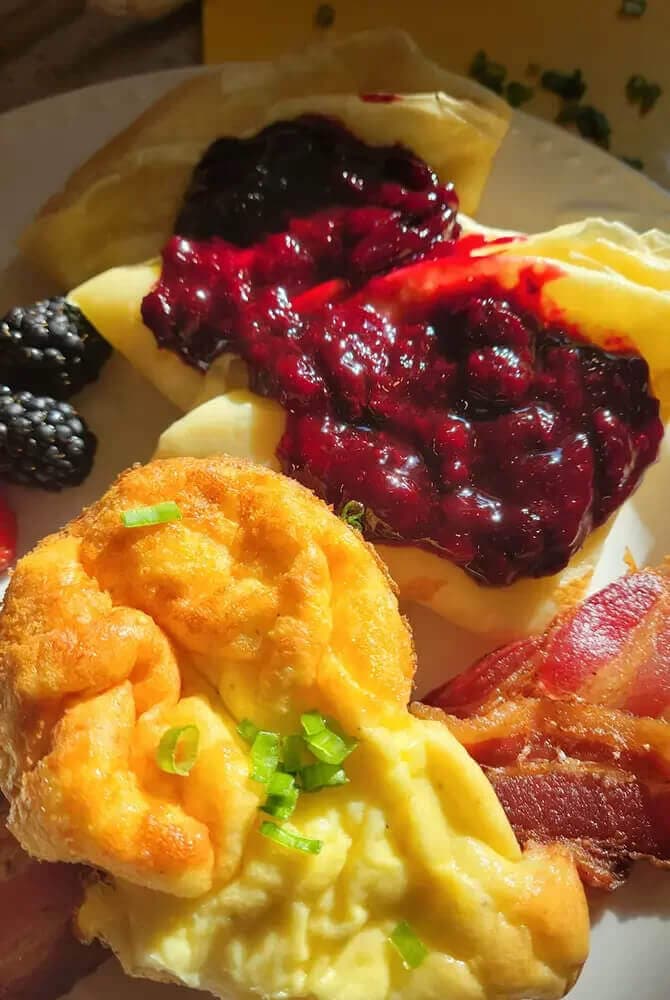 A plate of breakfast items including pancakes with berry sauce, a fluffy egg dish, bacon, and fresh blackberries.