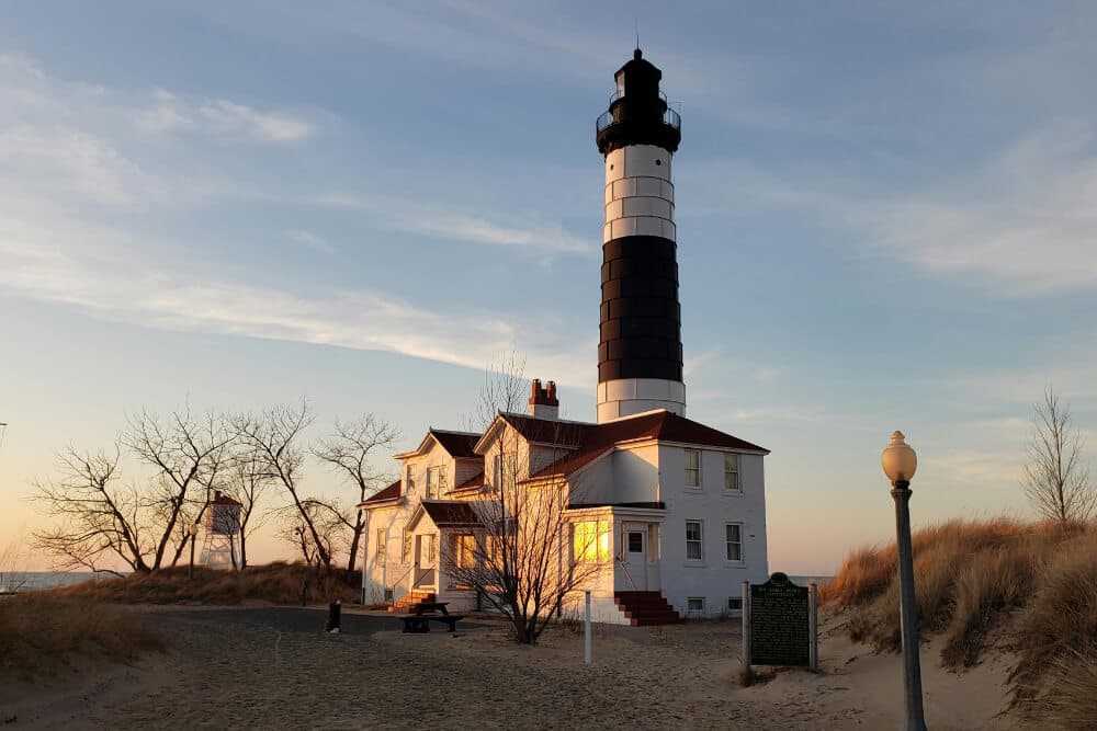 A black-and-white striped lighthouse stands beside a sandy beach at sunset, with a white house nearby.